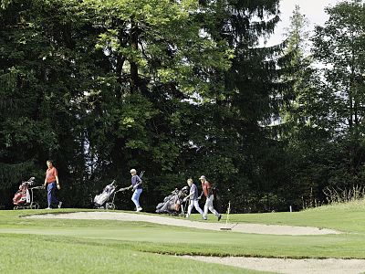 Golfers walk on a green path, carrying golf bags, with tall trees and greenery in the background, evoking a sense of calm and recreation.