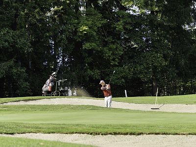 A golfer swings a club near a sand bunker on a green golf course surrounded by trees. A golf bag is visible in the background.