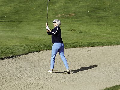An elderly person golfing in a sand trap, swinging a club with grassy surroundings.