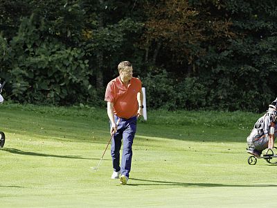 A man in a red shirt and blue pants plays golf on a green course, with trees in the background and two golf carts visible.