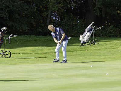 A man putting on a golf green, with two golf bags on wheels in the background on a sunny day. Trees outline the course behind him.