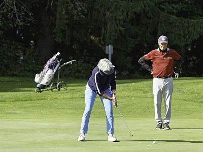 A golfer in blue jeans puts on a green lawn, while another person in red observes. Golf bags can be seen in the background, surrounded by trees.