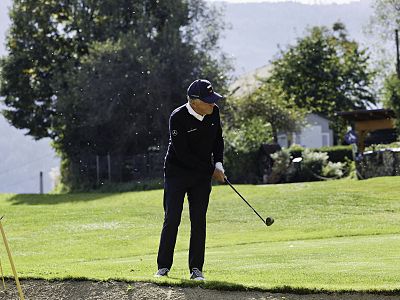 Golfer in black attire, ready to swing on a green course with trees and a house in the background under a clear sky.