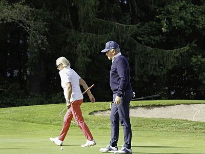 Zwei Personen beim Golfspielen auf einem grünen Golfplatz. Eine Person trägt rote Hosen und ein weißes Shirt, die andere ist dunkel gekleidet.