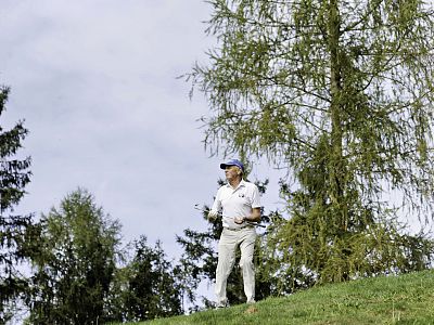 Man in white with golf clubs walks on a grassy hill, surrounded by trees under an overcast sky.