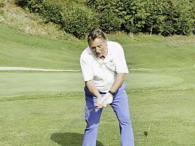 A man on a golf course swinging a club, wearing a white shirt and blue pants, surrounded by green grass and trees.