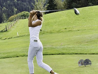 A golfer in striped pants swings a club on a verdant course under a clear sky, with hills and trees in the background.