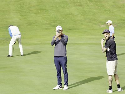 Two men stand on a golf course, one talking into a phone and the other smiling. A third golfer bends to line up a putt in the background.