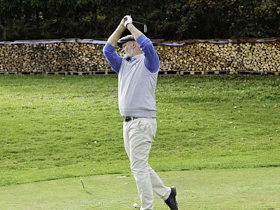 A golfer in a sweater and hat swings a club on a grassy course, with stacks of cut logs behind them.