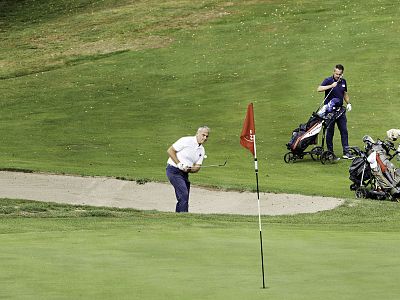 A golfer in white shirt and blue pants prepares to hit a ball from a sand trap. Another person stands nearby with golf equipment on a green course.
