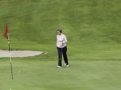 An elderly woman on a golf course near a flag, holding a club, attentively watching the golf ball on the green field.