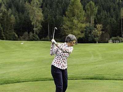 Woman golfing on a green course, mid-swing with a patterned shirt, surrounded by trees and grass.
