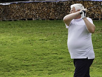 A man in a white shirt and black pants swings a golf club on a grassy field with wooden logs piled in the background.