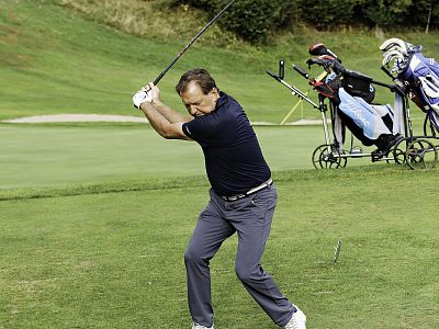 Man in navy shirt and gray pants swinging a golf club on a lush green course, with golf bags on carts nearby.
