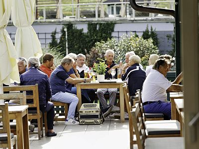 A group of elderly people sit around a wooden table at an outdoor café. They are chatting and enjoying drinks under large umbrellas on a sunny day.
