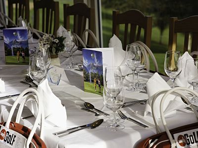 Elegant table setting with white napkins, wine glasses, and event brochures. Chairs and green scenery visible in the background, creating a warm atmosphere.