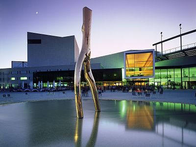 Modern sculpture on reflective surface, with illuminated contemporary buildings in the twilight.