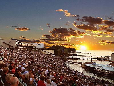Hundreds of people sit in an open-air amphitheater facing the sea. The sun is setting, casting a warm glow on the clouds and water, creating a peaceful scene.