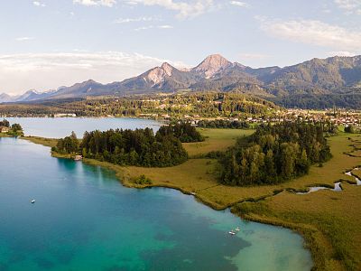 Aerial view of a clear blue lake surrounded by green fields and forests with mountains in the background under a partly cloudy sky.