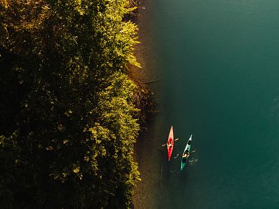 Aerial view of two kayakers on a blue lake near a dense forest, with vibrant red and green kayaks creating a striking visual contrast in the serene natural setting.