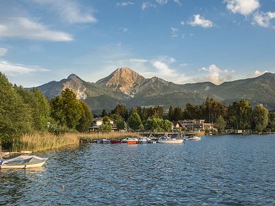 Serene lake with boats, surrounded by trees and houses, with mountains in the background and a clear, blue sky overhead.