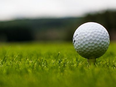Close-up of a golf ball on a tee in green grass, with a blurred background and trees in the distance, under a clear sky.