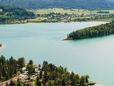 Aerial view of a serene lake with surrounding green landscape, small villages, and rolling hills. The water contrasts with the rich greenery around.
