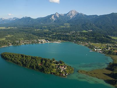 Aerial view of a turquoise lake surrounded by lush landscape, featuring an island and a background of tall, majestic mountains under a clear sky.