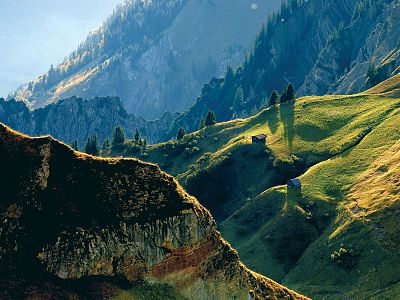 Bright green hills and rocky mountain peaks under sunlight, small cabin visible on hillside.
