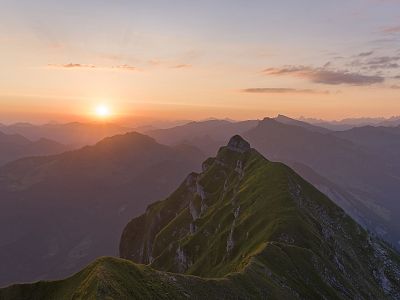 A majestic mountain range at sunset with orange and purple hues in the sky and a prominent jagged peak in the foreground.