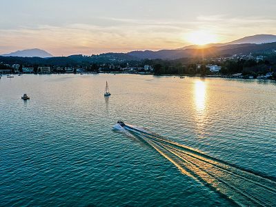 A scenic sunset over a tranquil lake with a boat creating ripples on the water. Mountains and a golden sky complete the peaceful vista.