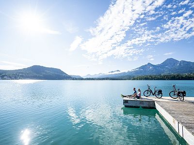 Two people sit on a lakeside pier under a clear sky, with bicycles nearby. Mountains and trees are reflected in the tranquil water.