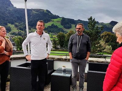Four people stand on a patio overlooking lush hills, engaged in conversation. They are casually dressed, with patio furniture visible around them.