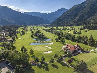 Vogelperspektive eines grünen Golfplatzes in einem Tal, umgeben von bewaldeten Hügeln und Bergen bei klarem Himmel. Ein Clubhaus ist rechts im Bild sichtbar.