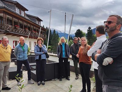 Eine Gruppe von Menschen steht draußen auf einer Terrasse vor einem Gebäude in den Bergen. Der Himmel ist bewölkt und die Landschaft wirkt kühl.