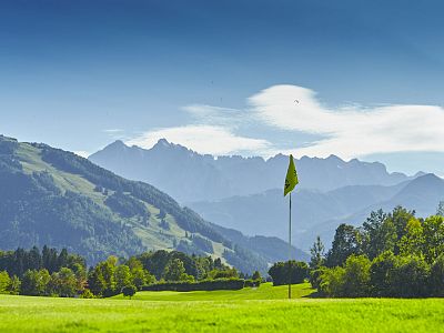 Golffeld in einer malerischen Berglandschaft, umgeben von grünen Wiesen und Bäumen. Ein gelbes Flaggenloch steht im Vordergrund, klare Himmel im Hintergrund.