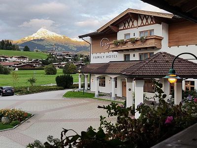 A family hotel in an alpine setting with a mountain in sunset light, lush greenery, and nearby parked cars.