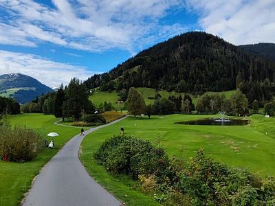 A scenic golf course with green hills, a curving path, and a pond under a blue sky with clouds.