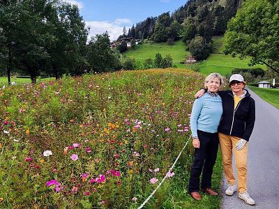 Two people stand on a path by a vibrant wildflower field, with lush green hills and trees in the background. The sky is partially cloudy.