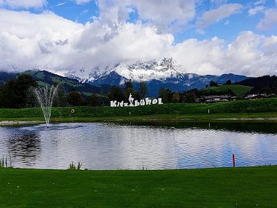 View of a lake with a fountain, mountains in the background, partially cloudy sky. Scenic landscape of Kitzbühel, Austria.