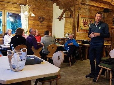 People sitting in a wooden cabin, talking and listening to a speaker. The setting is cozy with wooden furniture and decorative wall art.