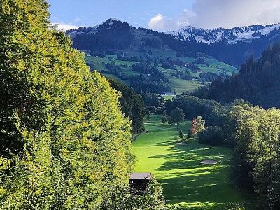 A lush green valley with a golf course, surrounded by trees and hills. Snow-capped mountains in the distance under a clear blue sky.