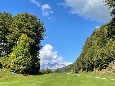 Green valley with trees on both sides and distant mountains under a blue sky with clouds.