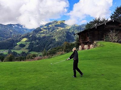 Person playing golf on a green field with mountains in the background, a wooden cabin nearby, under a partly cloudy sky.