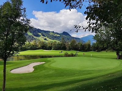 A scenic golf course with a green, sand trap, and trees, set against mountains and a blue sky with clouds.