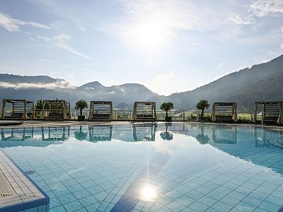 Outdoor pool reflecting mountains and sky, with sun loungers nearby and hills in the background. Clear, sunny blue sky and atmospheric tranquility.