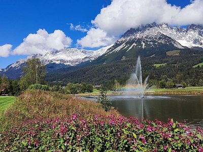 A lake with a fountain, colorful flowers, and snow-capped mountains under a blue sky.
