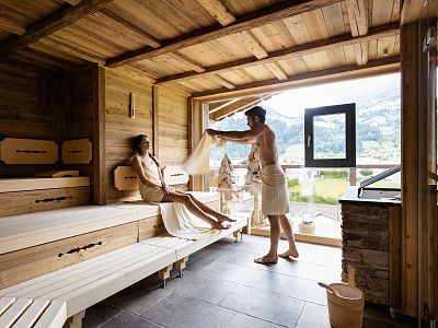 Two people relax in a wooden sauna with a scenic view through a large window. The room exudes warmth and tranquility, with wooden benches and bright lighting.