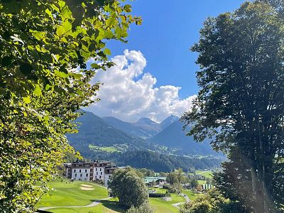 A scenic view of mountains and a small building with green hills and tall trees under a bright blue sky with white clouds in the Austrian Alps.