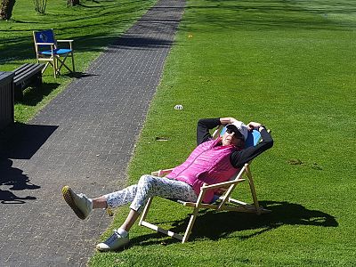Person in a pink vest reclines on a lounger on a grassy lawn under the sun, with a paved path and chair in the background.